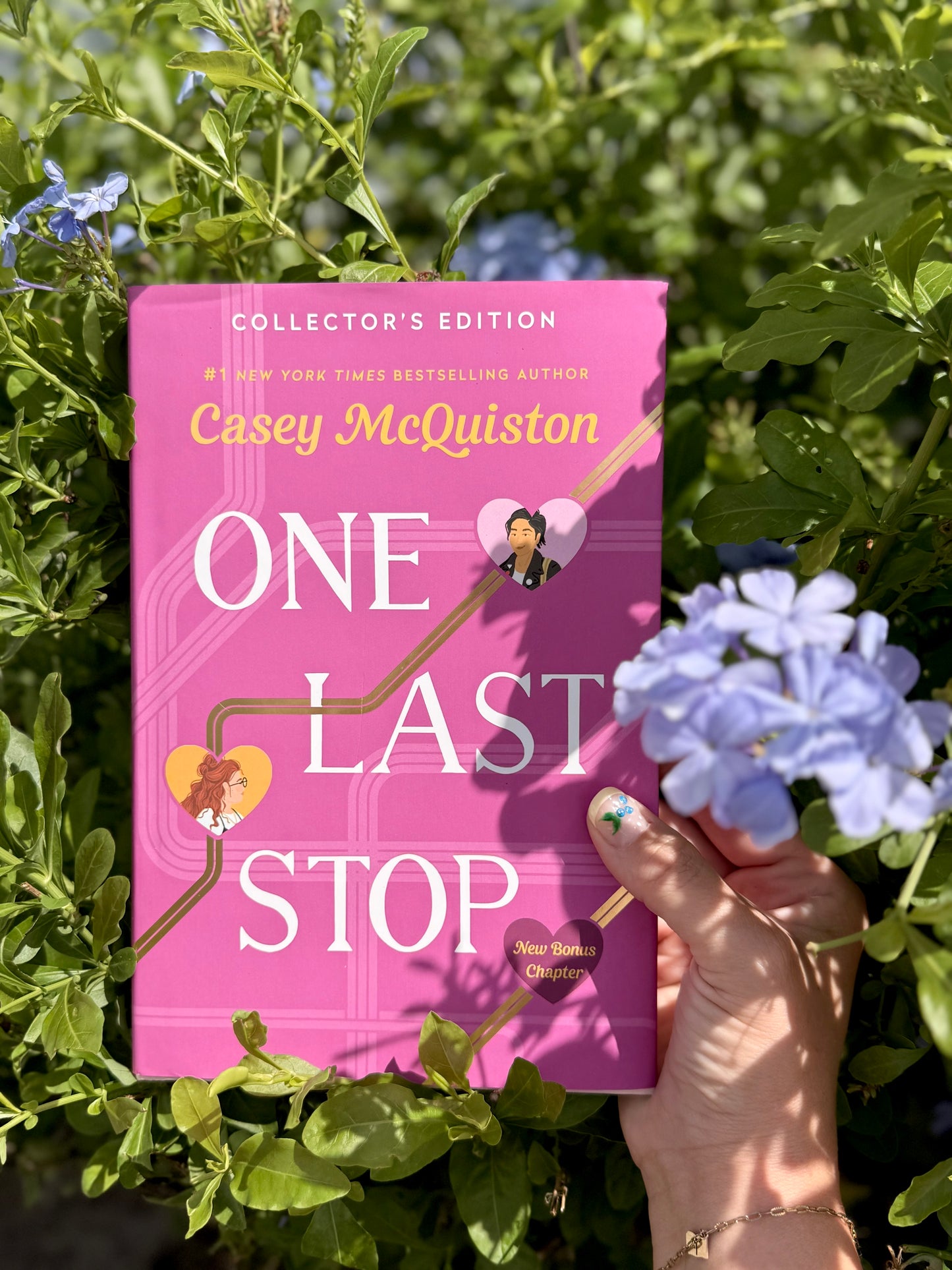 Pink book titled "One Last Stop" by Casey McQuiston. held by a feminine hand. Surrounded by greenery and blue flowers.